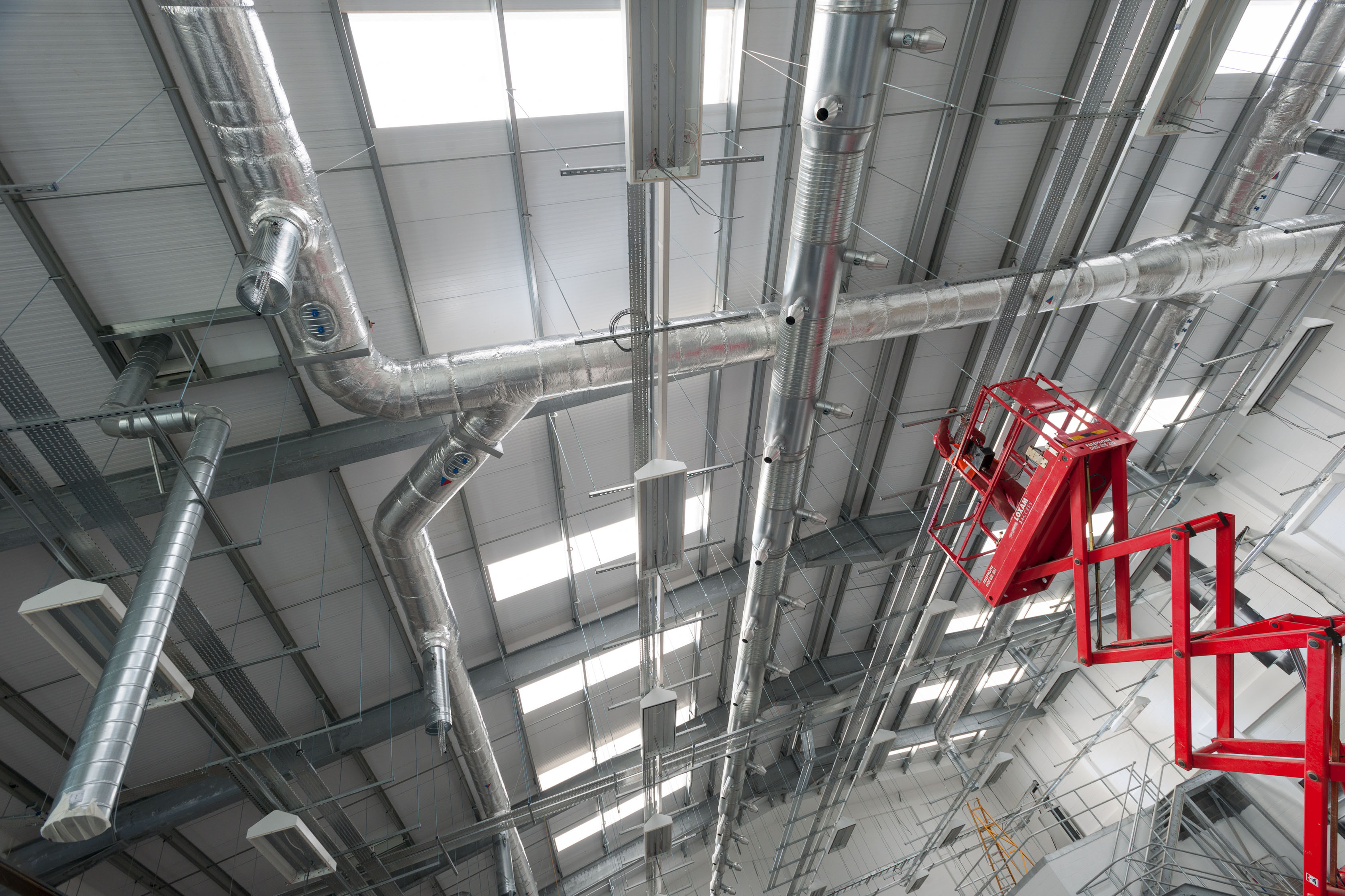 Man on red scaffolding tower fixing roof interior of silver grey industrial unit